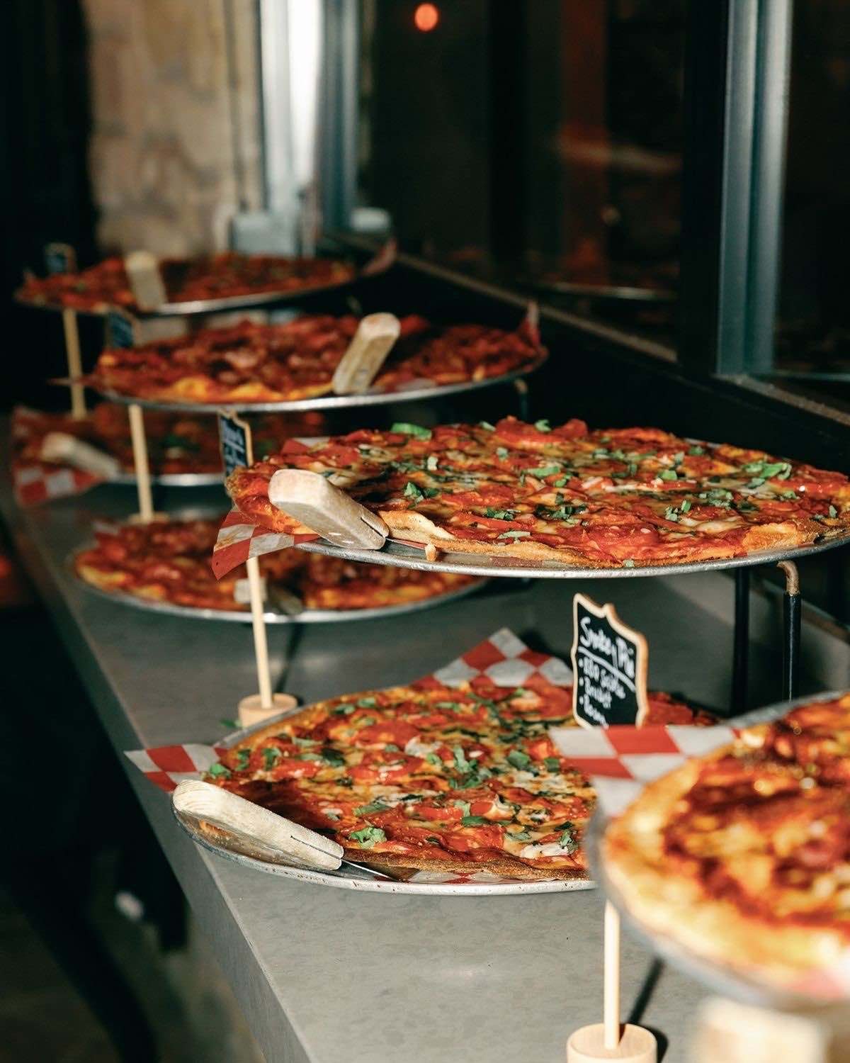 Mehrere große Pizzen auf Metalltabletts in Etagenständern auf einem Ausgabetresen, davor kleine Kreidetäfelchen mit den Sorten