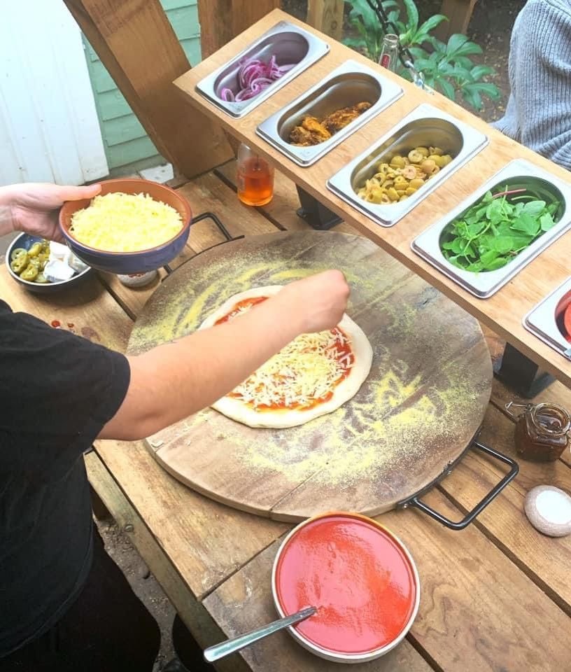 Pizzateig wird an einer Zubereitungsstation im Freien belegt, dahinter Behälter mit roten Zwiebeln, Oliven und Rucola