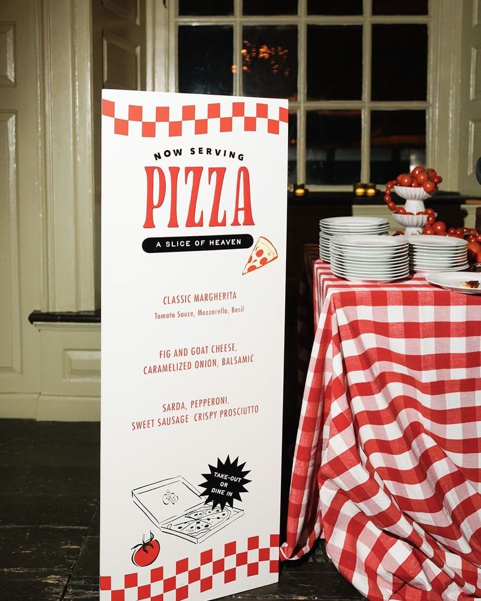 Aufsteller mit handgeschriebener Pizzakarte neben einem Tisch mit gestapelten Tellern und rot-weiß karierter Decke