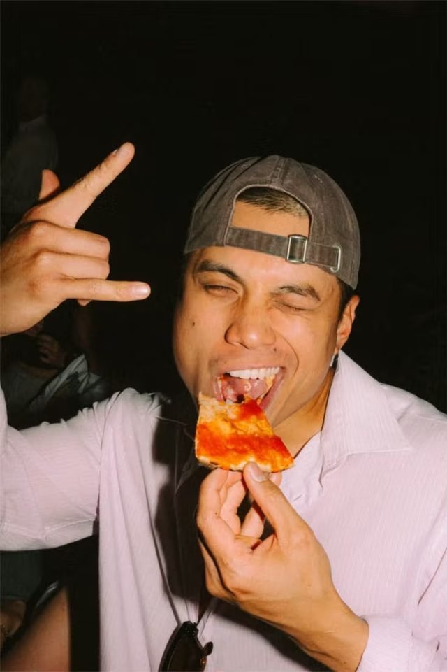 Ein Gast beißt lachend in ein Stück Pizza und reckt die freie Hand in die Höhe, Nachtszene