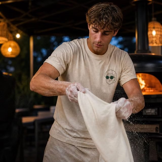Pizzabäcker im Nonna's-Crew-Shirt wischt sich die Hände am Tuch ab, im Hintergrund der Ofen