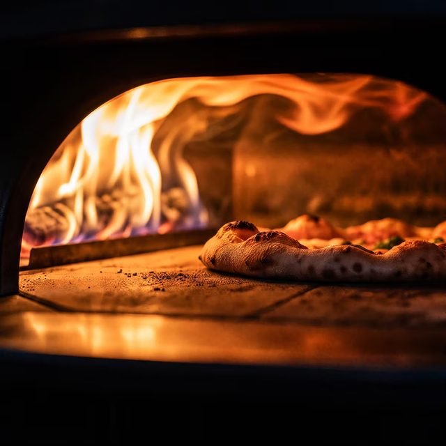 Blick in den Ofen: die Flamme züngelt an der Rückwand, davor bäckt eine Pizza auf dem Stein