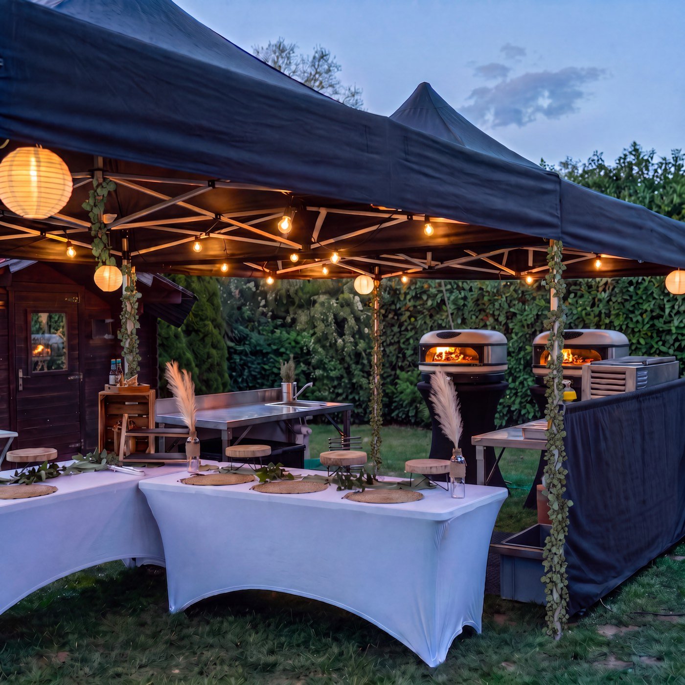 Aufgebauter Pavillon mit Lichterketten, eingedeckten Tischen und zwei Pizza-Öfen bei Dämmerung