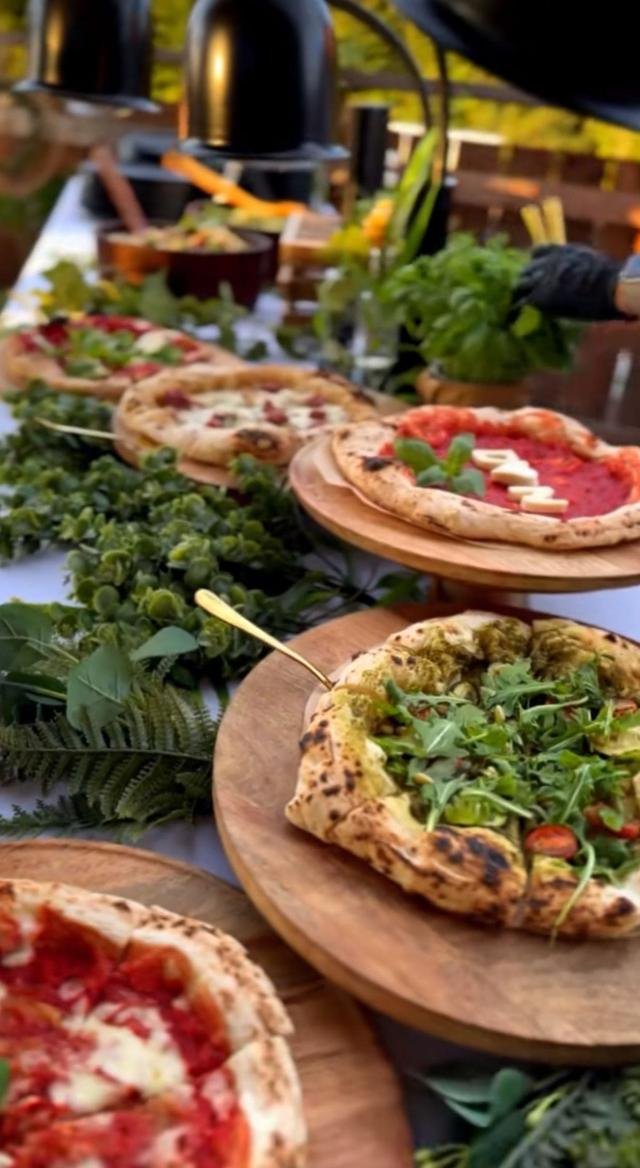 Frisch gebackene Pizzen auf erhöhten Holzbrettern zwischen grünen Kräutern, dahinter zwei Pizzaöfen