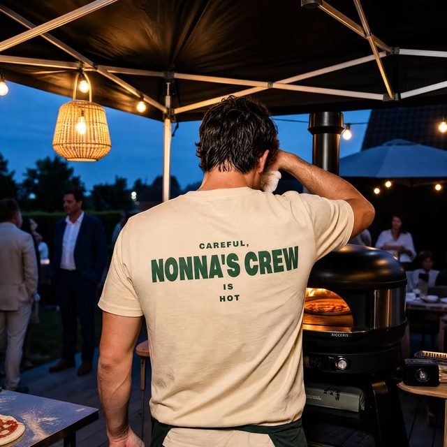 Person von hinten im hellen Nonna's-Crew-Shirt unter dem Pavillon, rechts daneben der Ofen bei Dämmerung
