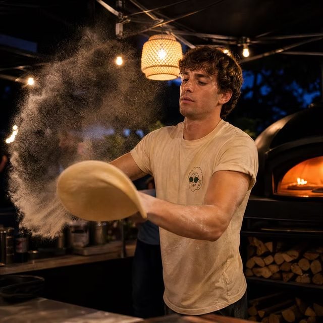 Ein Pizzabäcker schwingt den Teigfladen in die Länge, eine große Mehlwolke steht in der Luft, dahinter der glühende Ofen unter dem Pavillon