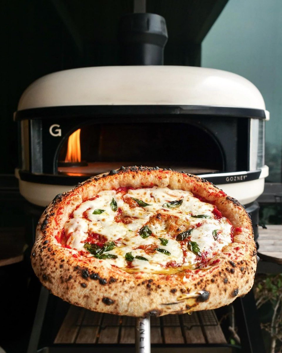 Eine Pizza auf der Schaufel vor einem hellen Gozney-Ofen mit brennender Flamme.